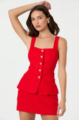 A woman is wearing a sleeveless, fitted red dress with wide shoulder straps. The dress features a square neckline and a front row of decorative gold buttons. The skirt portion of the dress is short and includes a layered, peplum-style overlay with two small flap pockets on the front.