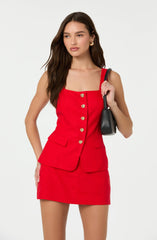 A woman is wearing a sleeveless red dress with a square neckline and gold decorative buttons down the front. The dress features two front flap pockets at the hips. She is carrying a small black handbag over her shoulder.