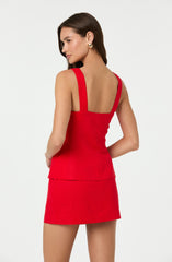 A woman is wearing a sleeveless, fitted red dress with wide shoulder straps. The top part of the dress is tailored and slightly peplum-style, with a layered design at the waist. The skirt is straight and short, ending above the knees. She accessorizes with gold hoop earrings.