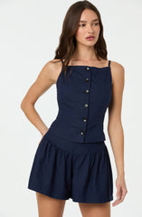 A young woman is wearing a sleeveless navy blue top with thin shoulder straps and a row of black buttons down the front. She pairs it with matching navy blue high-waisted shorts that have a gathered, loose-fitting design.