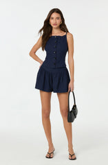 A woman is wearing a navy blue sleeveless top with thin spaghetti straps and a row of black buttons down the front. She is also wearing matching navy blue shorts with an elastic waistband and gathered fabric. She has on black strappy high-heeled sandals and is carrying a small black handbag with a top handle. She has long, wavy brown hair and is posing with one hand on her hip against a plain light-colored background.