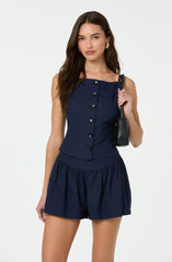 A woman is wearing a sleeveless navy blue top with a row of black buttons down the front and matching navy blue shorts with an elastic waistband and gathered fabric. She is holding a black shoulder bag and has long, wavy brown hair. She is also wearing gold earrings.