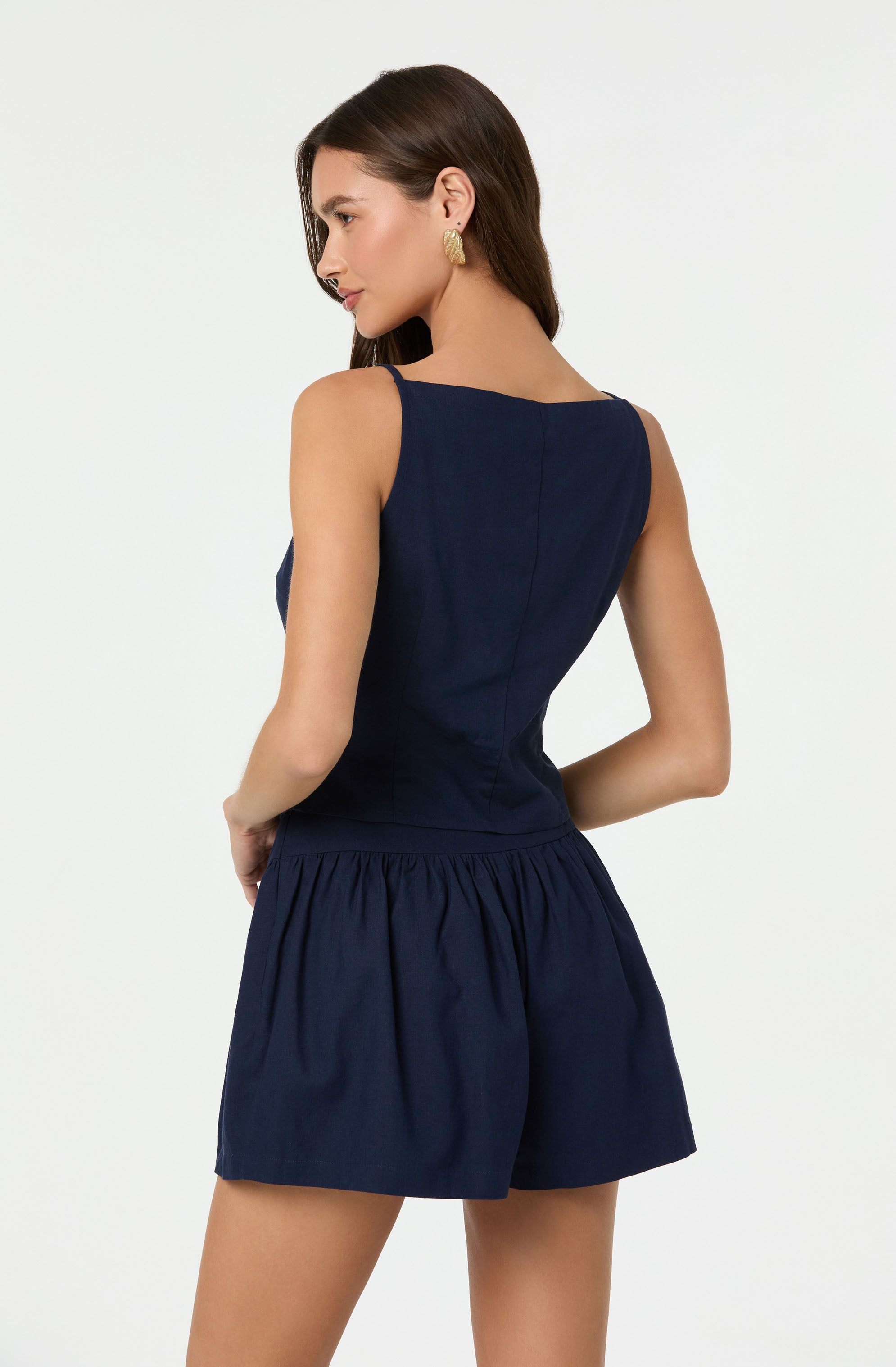 A woman is wearing a sleeveless, dark navy blue top with a fitted waist and matching navy blue shorts with an elastic waistband and gathered fabric. She has long, dark hair and is accessorized with gold earrings.