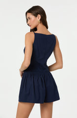 A woman is wearing a sleeveless, dark navy blue top with a fitted waist and matching navy blue shorts with an elastic waistband and gathered fabric. She has long, dark hair and is accessorized with gold earrings.