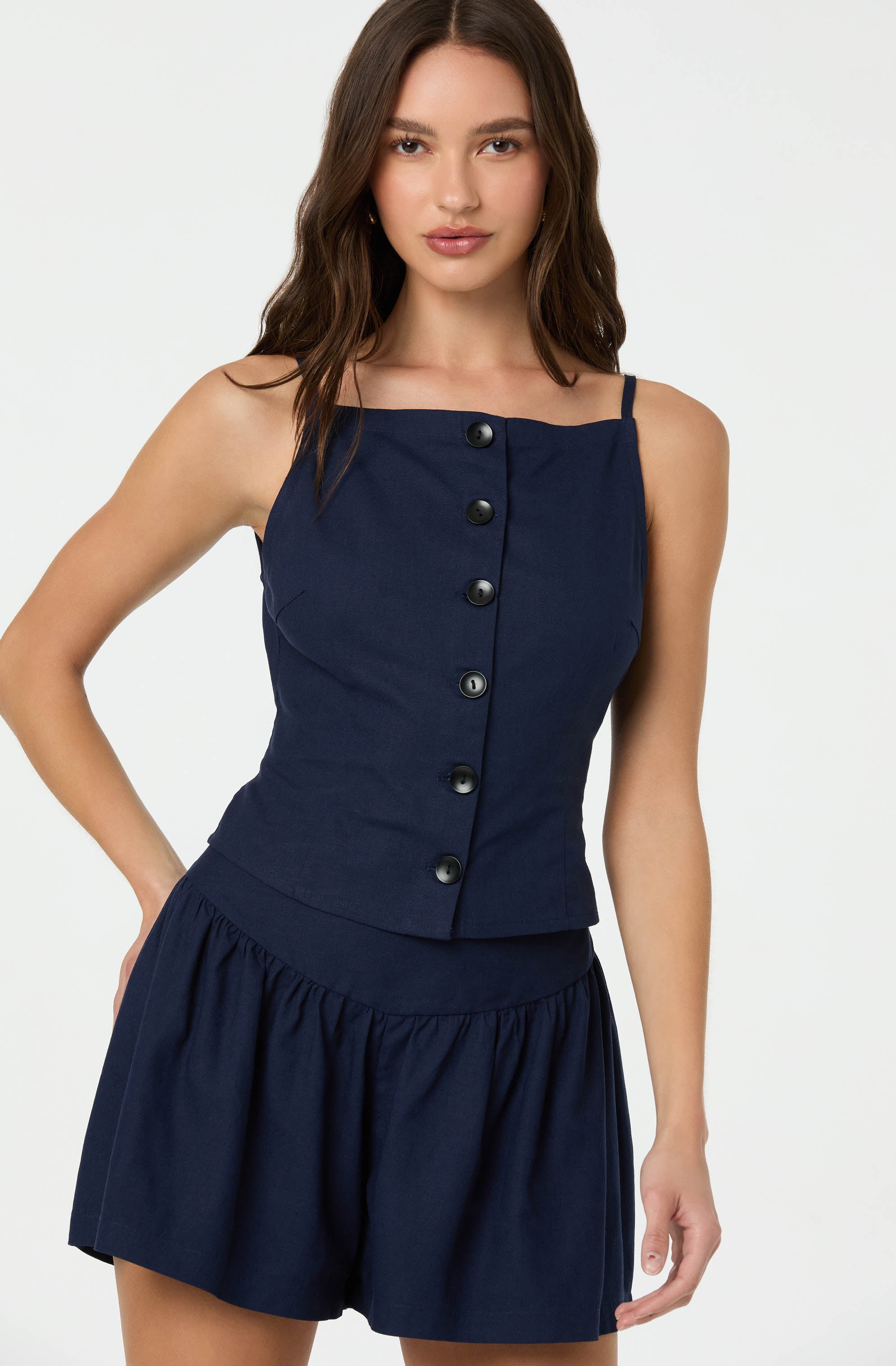 A woman is wearing a navy blue sleeveless top with thin straps and a straight neckline, featuring a row of black buttons running down the front. She pairs it with matching navy blue shorts that have a gathered, elastic waistband and a loose, flowy fit.