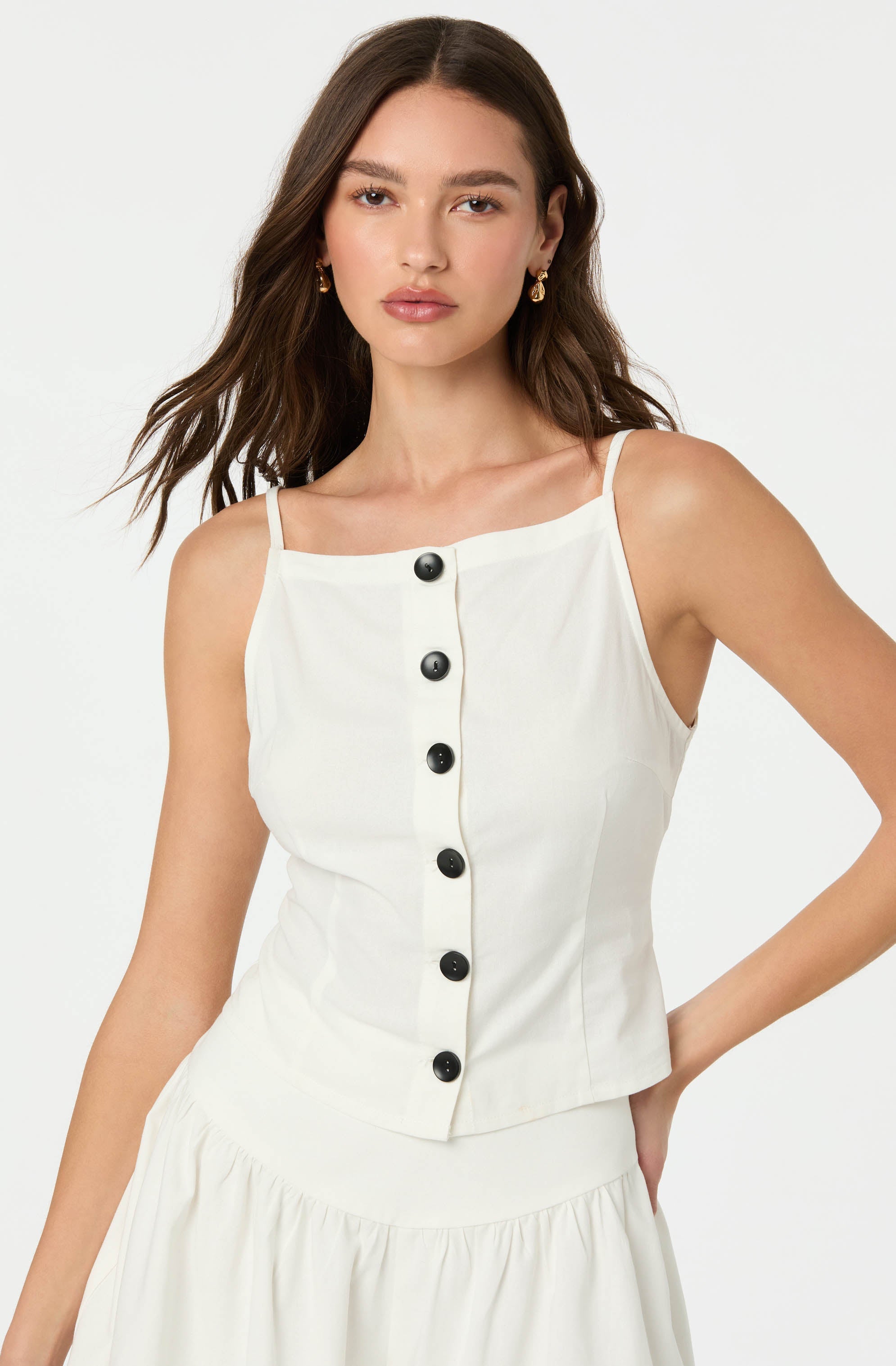 A woman is wearing a sleeveless, cream-colored top with thin spaghetti straps and a front row of black buttons. The top has a fitted silhouette and is paired with a matching cream skirt with a gathered, ruffled detail at the waist.