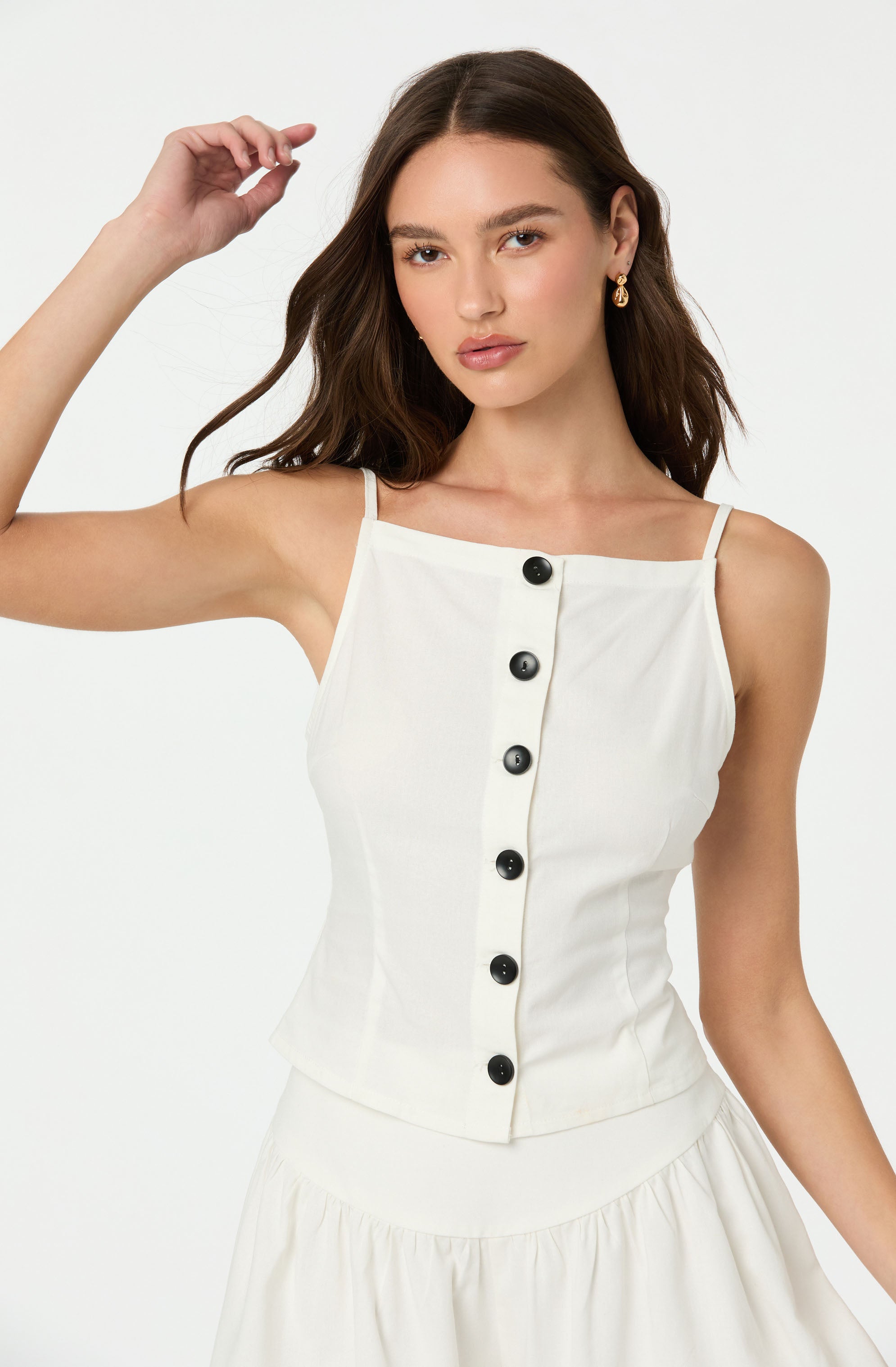 A young woman with long, wavy brown hair wearing a sleeveless white top with thin spaghetti straps. The top features a center row of large black buttons running vertically down the front. She is also wearing a pair of gold earrings.