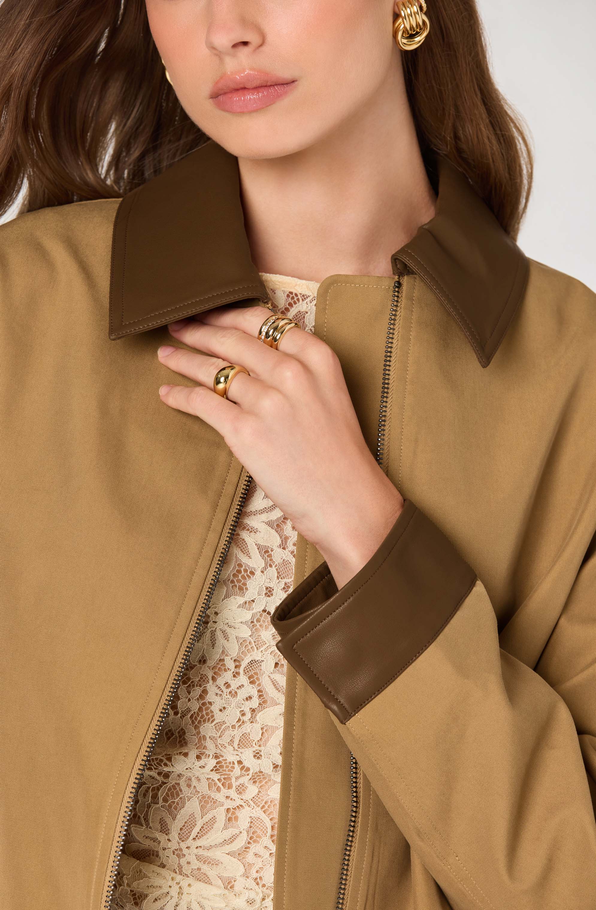 A woman is wearing a tan jacket with a dark brown collar and cuffs. The jacket is partially unzipped, revealing a light-colored lace top underneath. She has gold jewelry, including large hoop earrings and multiple rings on her fingers.