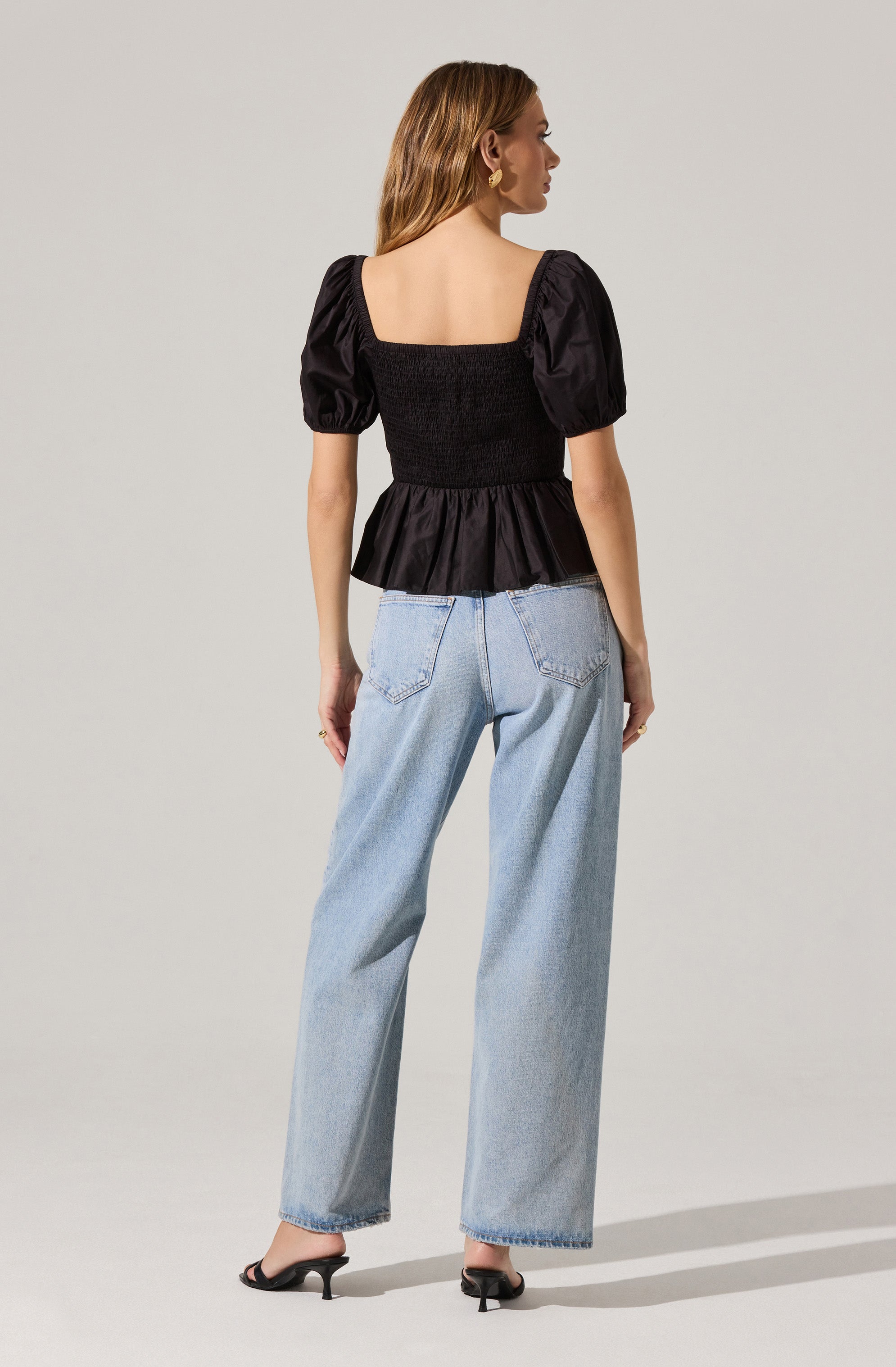 A woman is wearing a black blouse with puffed short sleeves, a smocked bodice, and a ruffled hem. She is also wearing light blue, high-waisted, wide-leg jeans and black heeled sandals. She has with gold hoop earrings and a ring on her right hand.