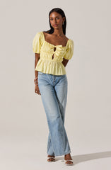 A woman is wearing a light yellow, short-sleeved blouse with puffed sleeves and a square neckline. The blouse features gathered fabric with two front keyhole cutouts, tied with bows, and has a ruffled hem. She pairs it with light-wash, high-waisted, wide-leg jeans that reach the floor. She is also wearing open-toe heels.