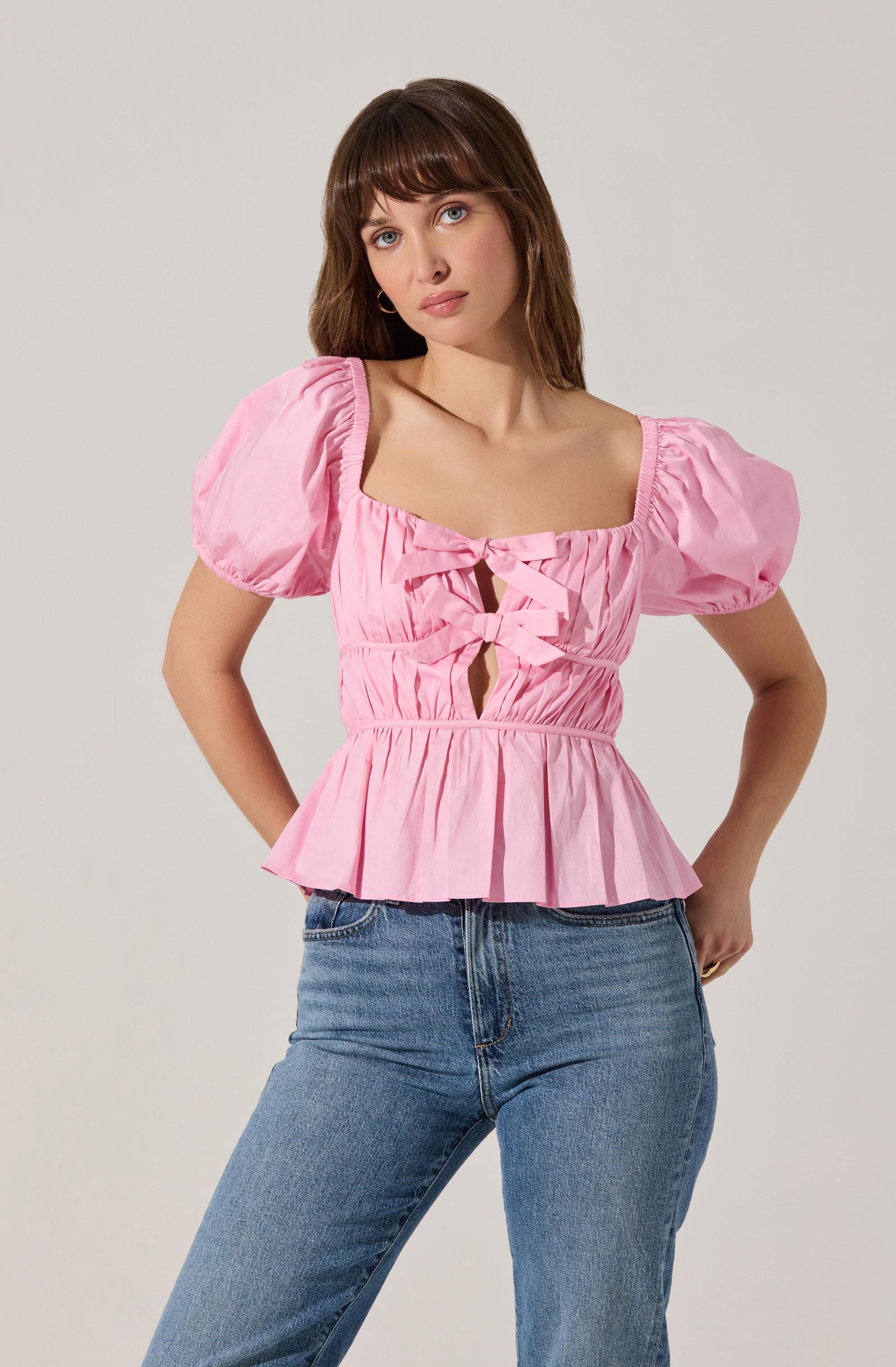 A woman is wearing a pink, short-sleeved blouse with puffed sleeves and a square neckline. The blouse features ruched detailing, decorative bow accents on the front, and a ruffled hem. She pairs it with light-wash blue jeans.
