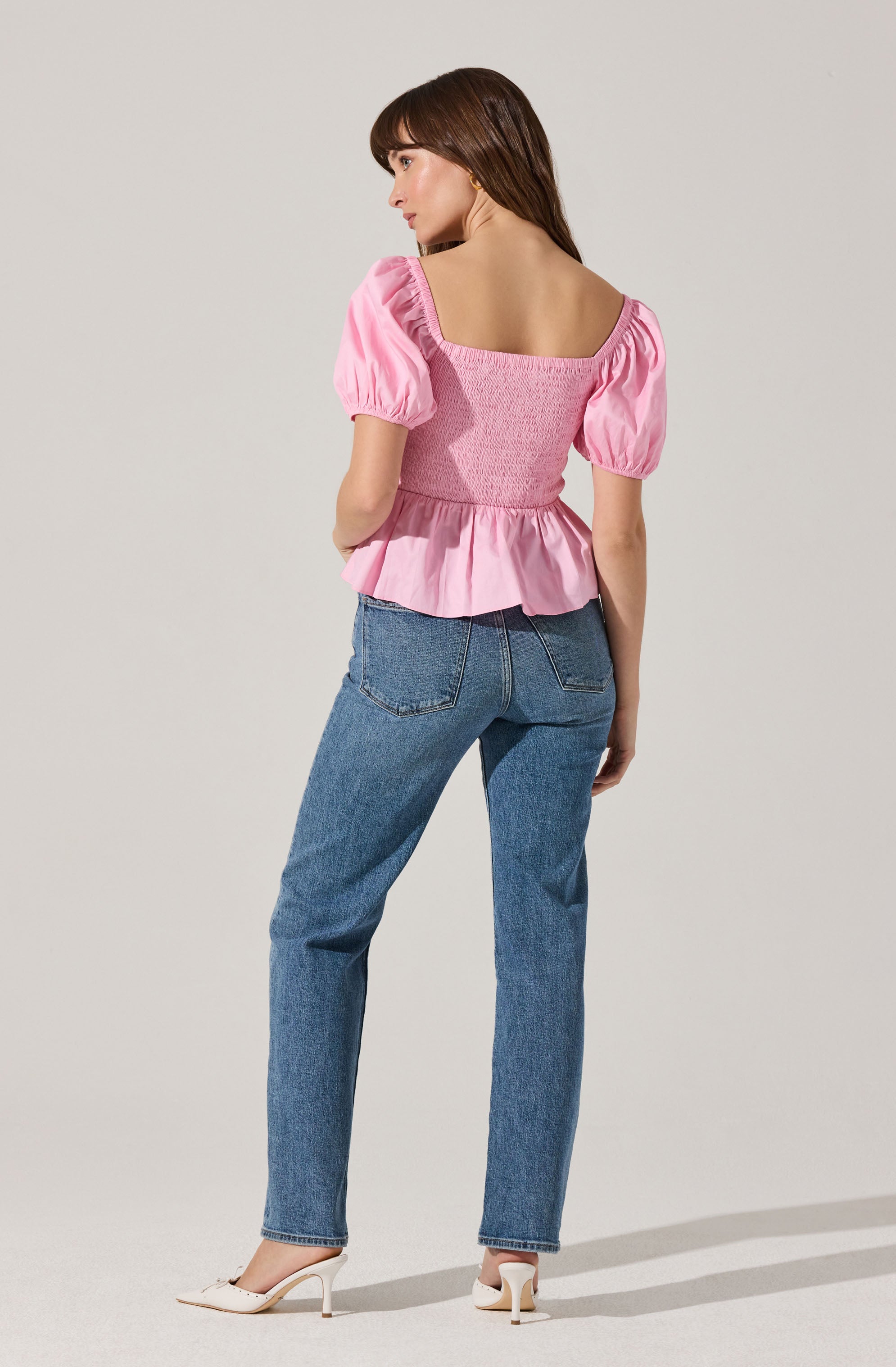 A woman is wearing a pink top with puffed short sleeves, a square neckline, and a ruffled hem that flares out at the waist. She pairs it with high-waisted blue jeans and white high-heeled shoes.