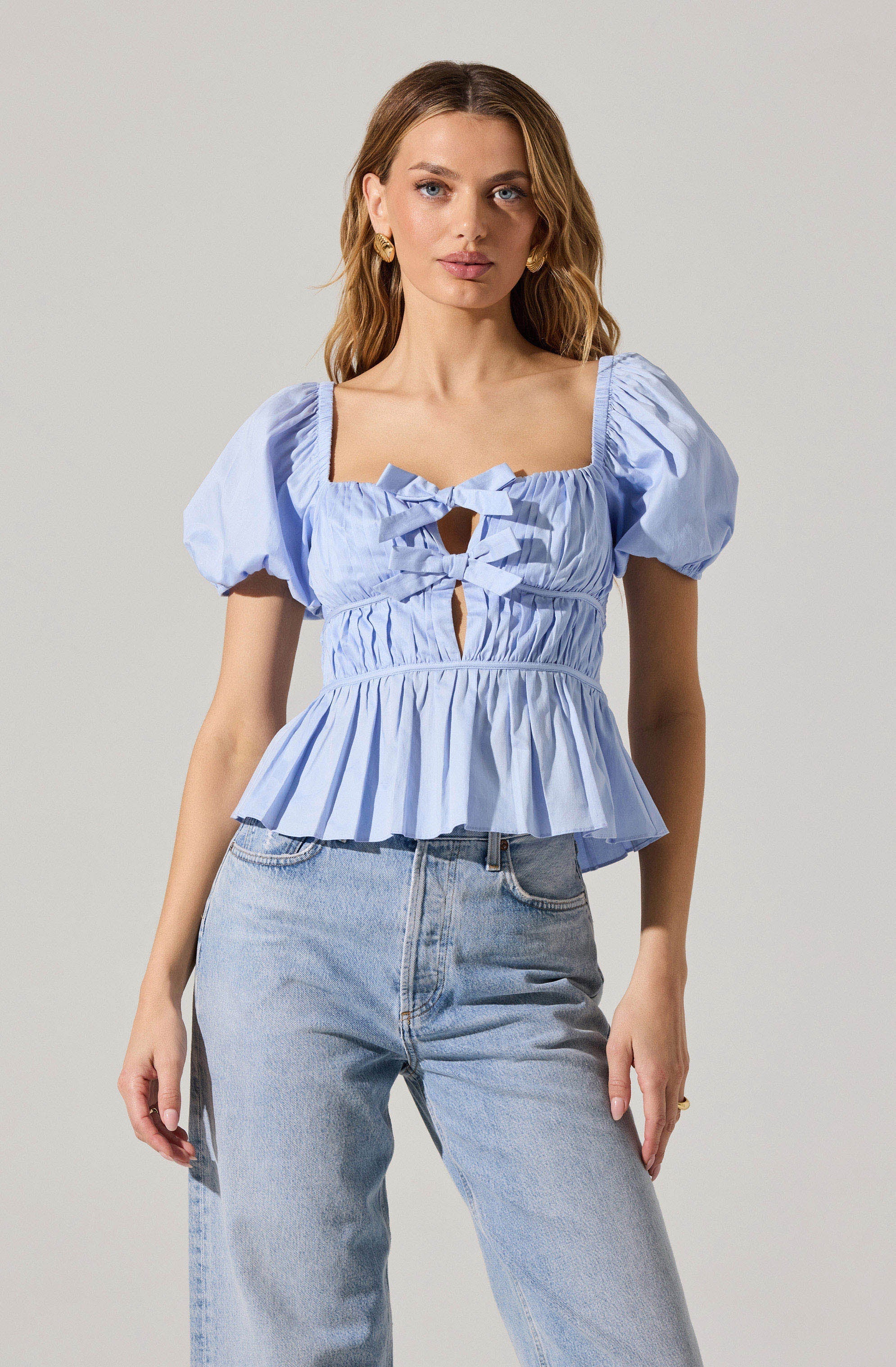 A woman is wearing a light blue, short-sleeved blouse with puffed sleeves and a square neckline. The blouse features ruched detailing, a front cut-out with tied bows, and a peplum hem.