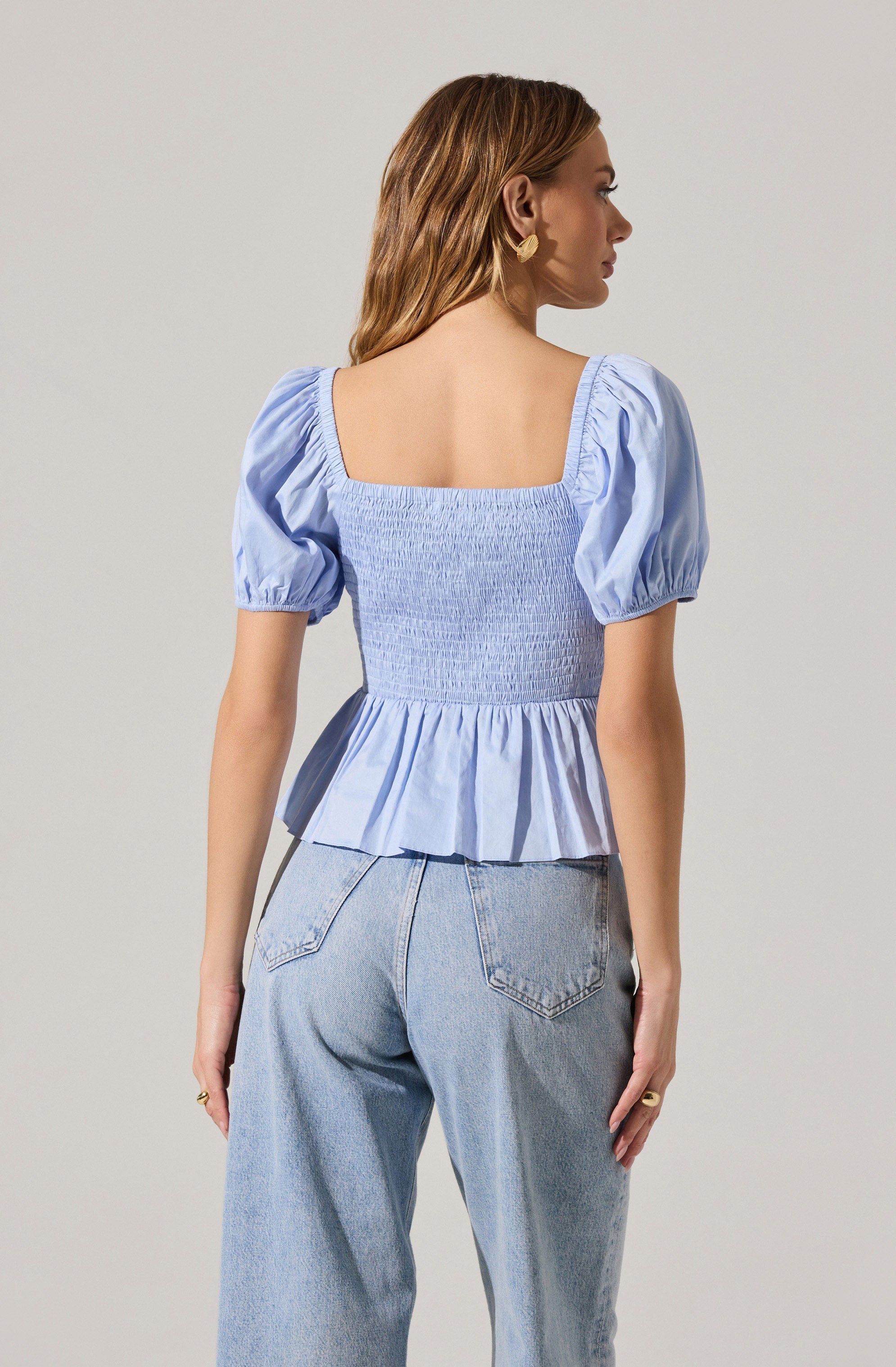 A woman is wearing a light blue, square-neck blouse with puffed short sleeves that have elastic gather at the edges. The blouse features a shirred, fitted bodice with a ruffled peplum hem. She pairs it with high-waisted, light-wash denim jeans.