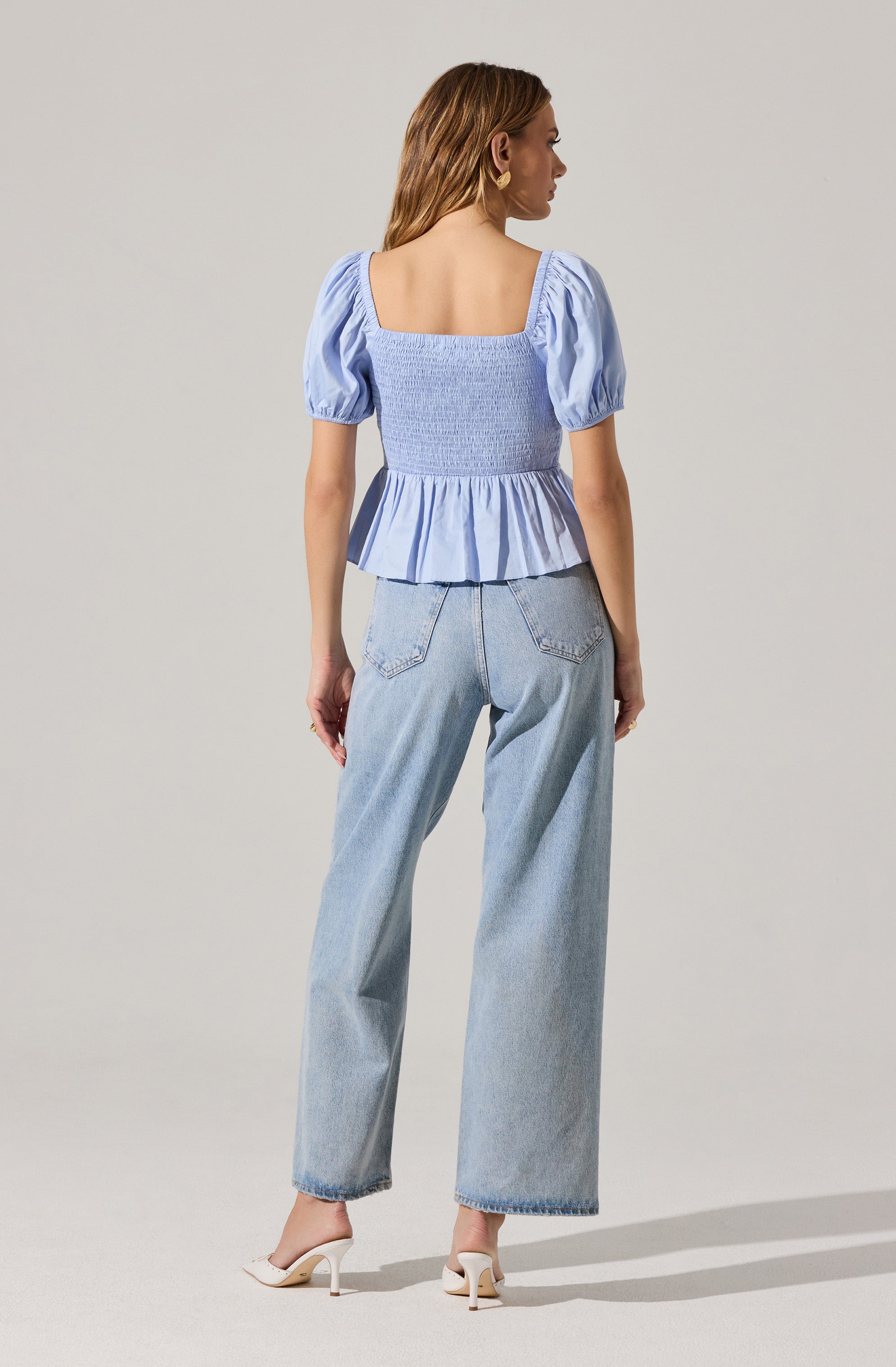 A woman is wearing a light blue, short-sleeved, square-necked top with puffed sleeves and a ruffled hem. The top features a smocked, fitted bodice. She is also wearing light-wash, high-waisted, wide-leg jeans and white high-heeled shoes.