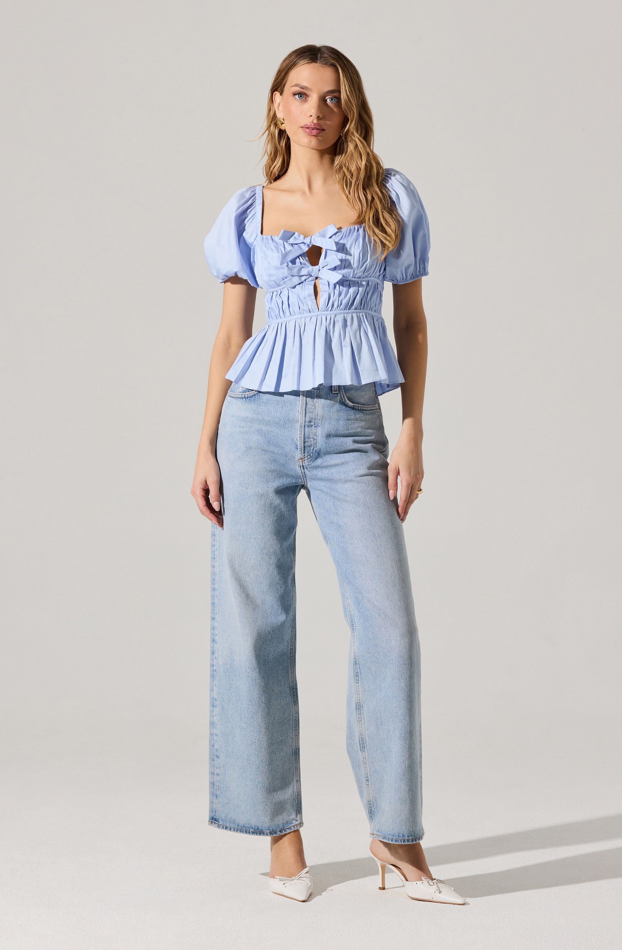 A woman is wearing a light blue, short-sleeved blouse with puffed sleeves and a square neckline. The blouse features a fitted bodice with ruffle details, cut-out sections in the front, and a peplum hem that flares at the waist. She pairs it with high-waisted, light-wash, wide-leg jeans and white high-heeled shoes.