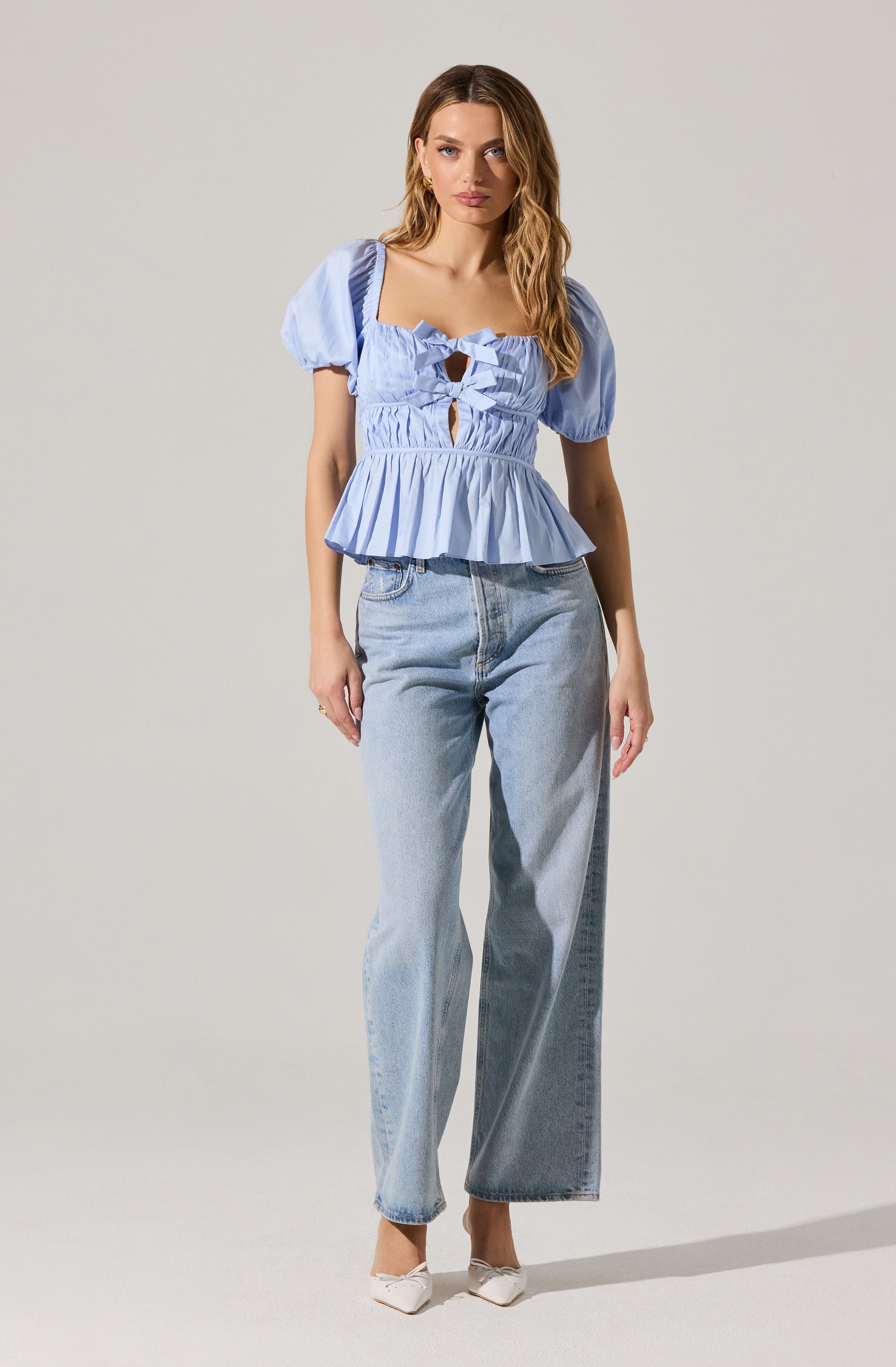 A woman is wearing a light blue puff-sleeve top with a square neckline, featuring horizontal ruffles and cutout details with bow accents on the front. The top has a peplum hem that flares out at the waist. She pairs it with high-waisted, loose-fitting, light-wash denim jeans and white pointed-toe shoes.