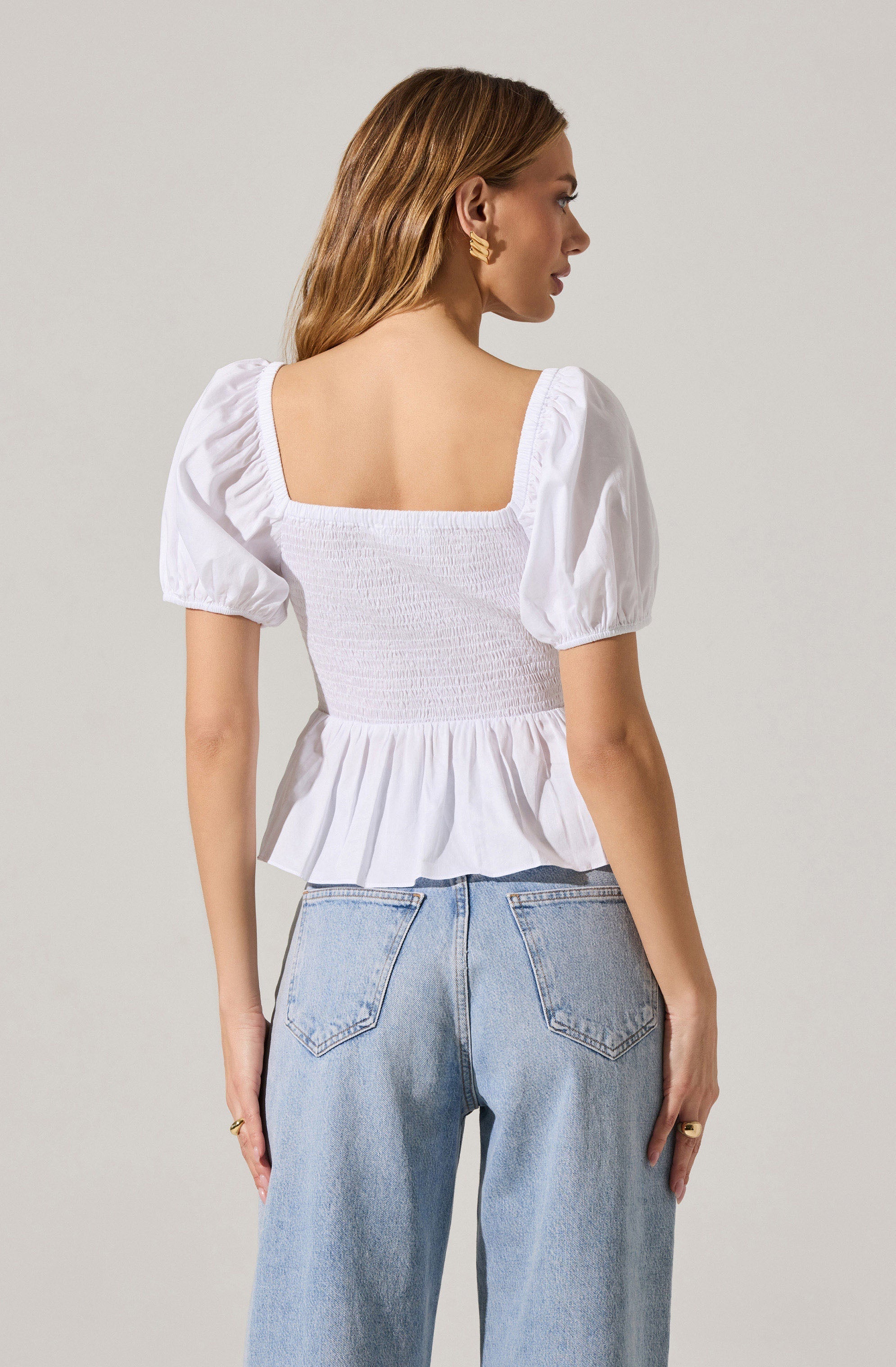 A woman is wearing a white top with a square neckline, puffed short sleeves, and a gathered, elasticized, ruched bodice with a ruffled hem. She is also wearing light blue high-waisted jeans.