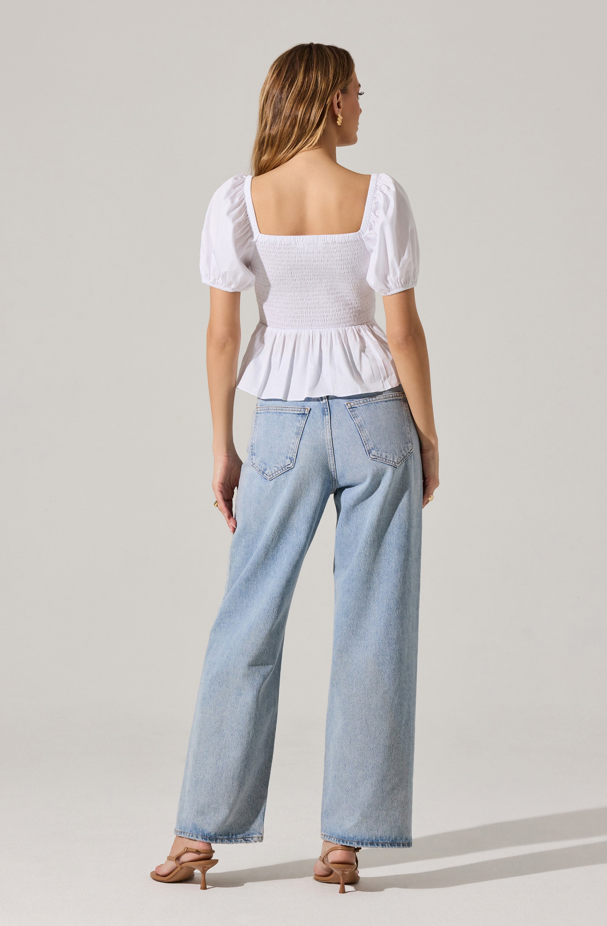 A woman is wearing a white, short-sleeved, smocked top with a square neckline and puffed sleeves, paired with light-wash, high-waisted, wide-leg jeans.