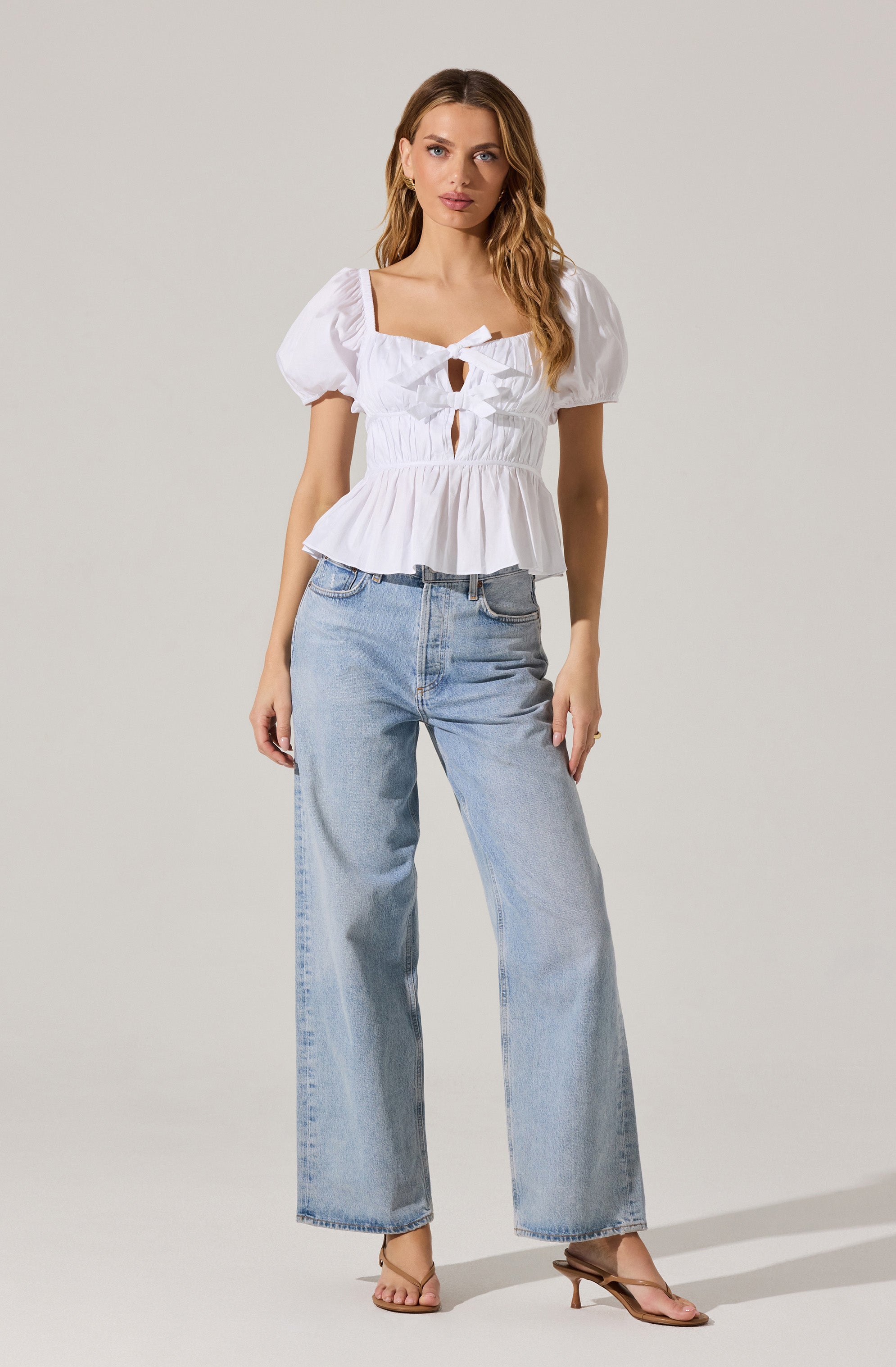 A woman is wearing a white blouse with puffed short sleeves, a square neckline, and a fitted bodice with gathered detailing and keyhole cutouts in the front. The blouse has a peplum-style hem that flares out at the waist. She pairs it with light-wash, high-waisted, wide-leg jeans and is wearing beige open-toe heels.