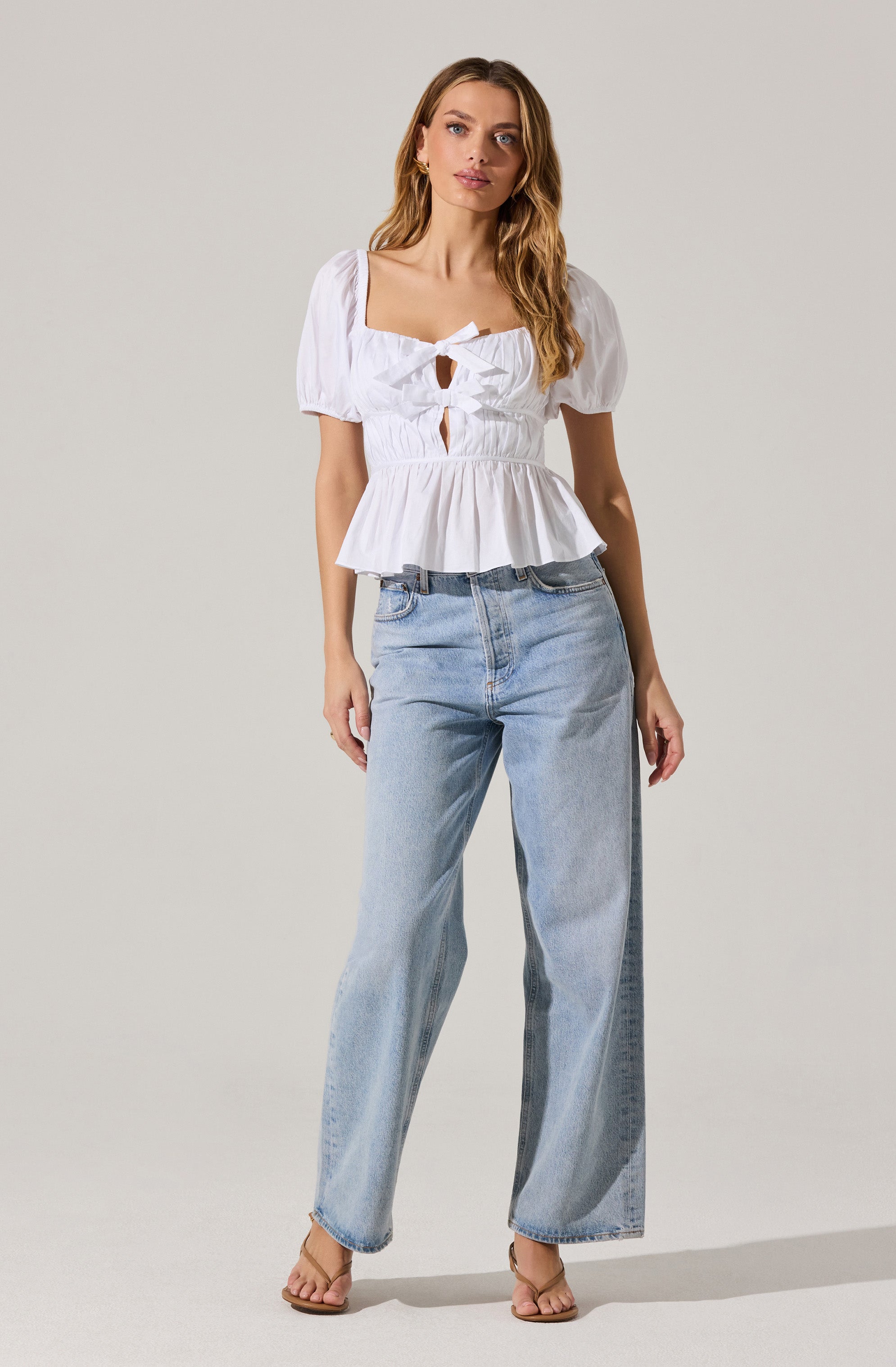 A woman is wearing a white, short-sleeved blouse with puffed sleeves and a square neckline. The blouse features front cutouts and a bow detail at the center, with a ruffled hem. She is also wearing light-wash, wide-leg jeans and tan sandals.