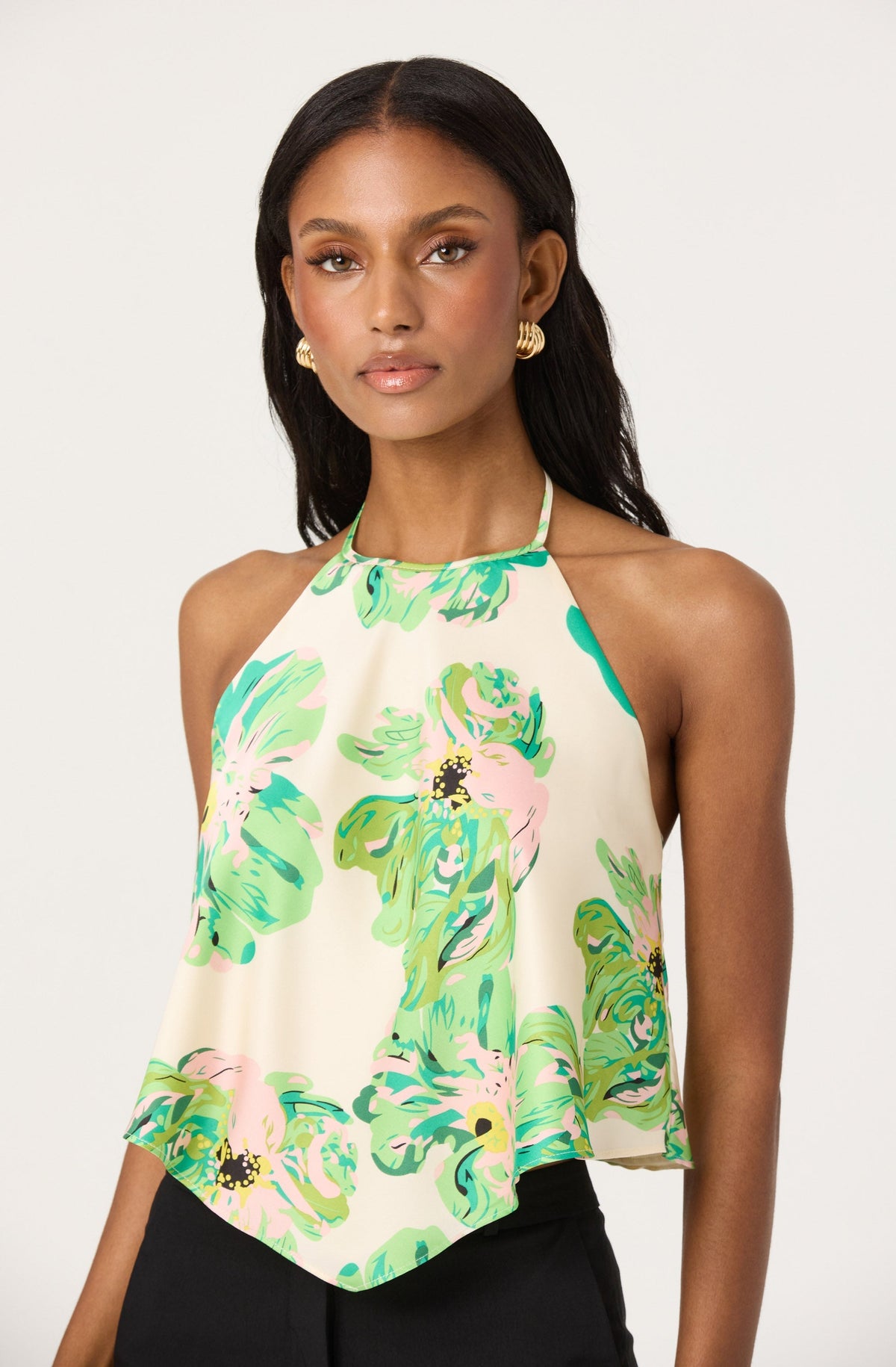 A woman is wearing a sleeveless halter top with a loose, flowy fit. The top features a high neckline with thin straps and is decorated with a colorful floral pattern in shades of green, pink, and yellow on a light cream background.