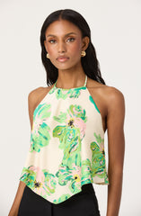 A woman is wearing a sleeveless halter top with a loose, flowy fit. The top features a high neckline with thin straps and is decorated with a colorful floral pattern in shades of green, pink, and yellow on a light cream background.
