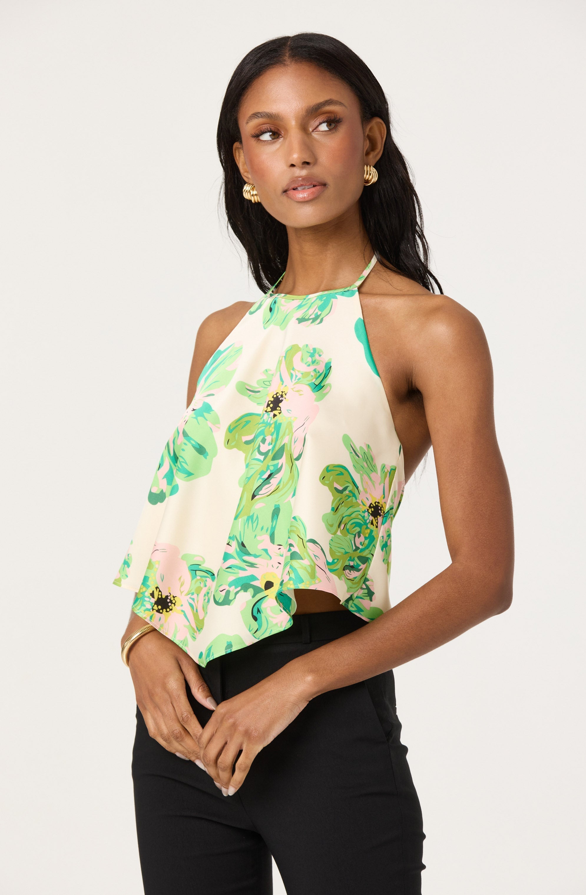 A woman is wearing a sleeveless halter top with a high neckline. The top features a colorful, abstract floral pattern in shades of green, pink, and black on a pale cream background. The fabric appears lightweight and flowy, draping loosely over her torso. She pairs the top with high-waisted black pants.