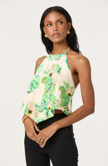 A woman is wearing a sleeveless halter top with a high neckline. The top features a colorful, abstract floral pattern in shades of green, pink, and black on a pale cream background. The fabric appears lightweight and flowy, draping loosely over her torso. She pairs the top with high-waisted black pants.