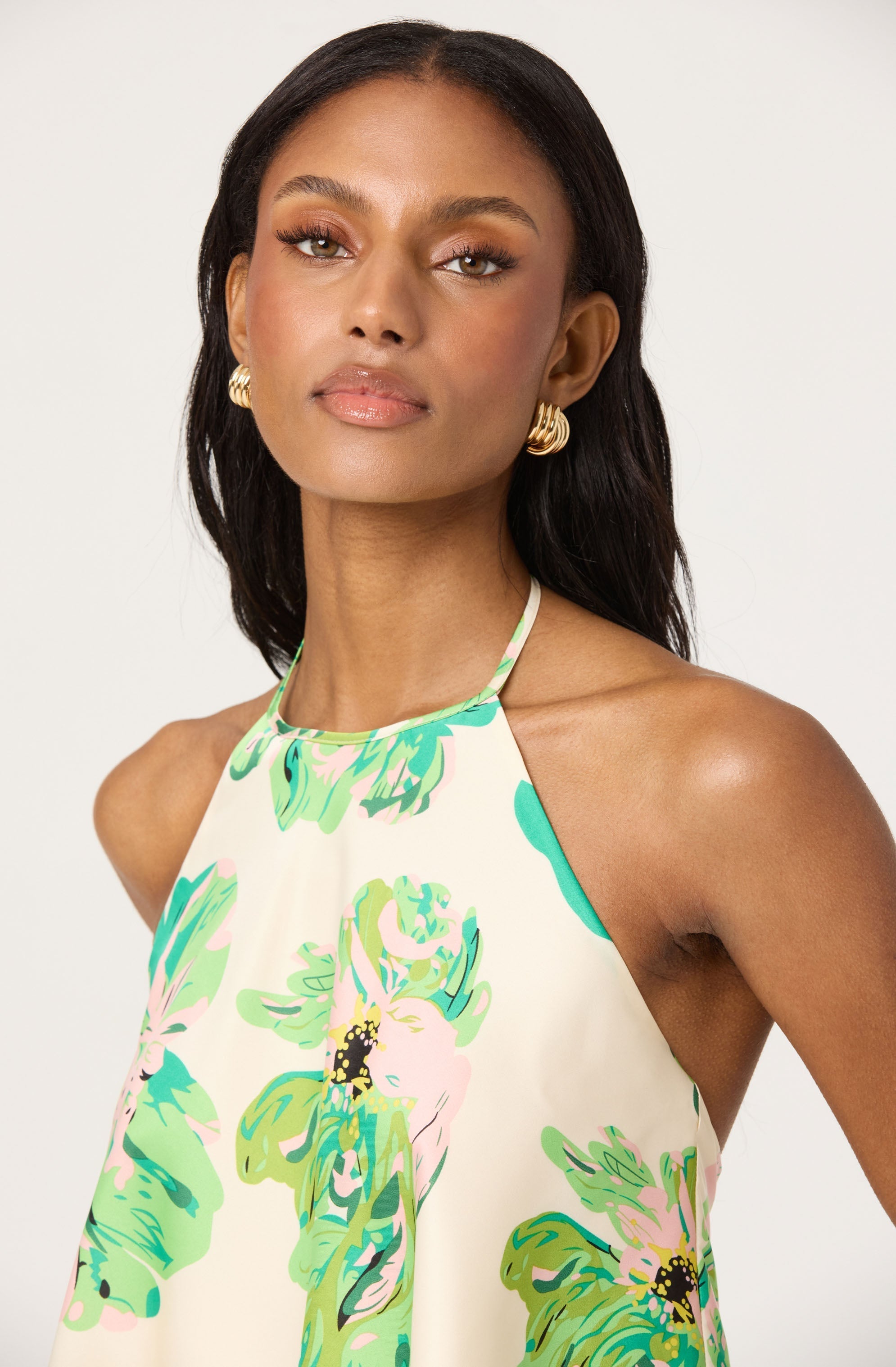 A woman is wearing a sleeveless, halter-neck dress with a floral pattern in shades of green, pink, and black on a light-colored background.