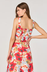A woman is wearing a sleeveless crop top and a matching high-waisted skirt, both made of fabric with a colorful floral pattern. The top features wide shoulder straps and a smocked, elasticized back. The skirt has a flowy fit, and both garments display a vibrant mix of red, white, light blue, orange, and green flowers.
