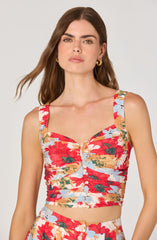 A woman is wearing a sleeveless, fitted crop top with wide straps. The top features a colorful floral pattern with large red flowers, green leaves, and light blue accents. The top has a button-down front with small white buttons running down the center.