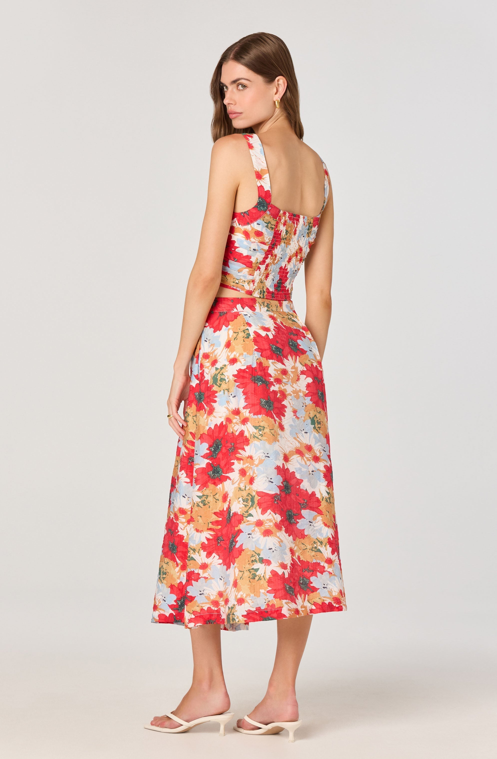 A woman is wearing a sleeveless, fitted crop top and a matching high-waisted, ankle-length skirt. Both pieces feature a vibrant, multicolored floral pattern with shades of red, pink, orange, yellow, and light blue. She is also wearing light-colored, open-toe high-heeled sandals.