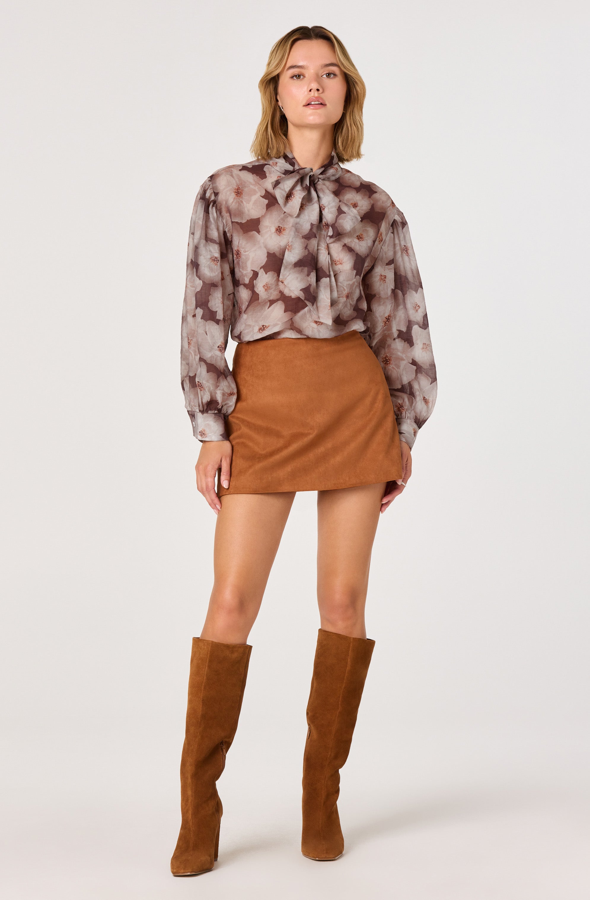 A woman is wearing a semi-sheer blouse with a floral print and a large bow at the collar, along with a high-waisted, tan suede mini skirt. She completes her outfit with knee-high tan suede boots.