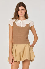 The outfit features a white short-sleeved blouse with a pointed, scalloped collar and a button-down front. Over the blouse, there is a fitted, sleeveless, light brown knitted top with a textured pattern. The bottom part of the outfit consists of beige, loose-fitting, pleated shorts with a gathered, bubble-style silhouette. The overall look combines soft tones and different textures.