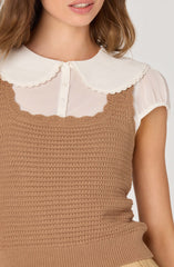 A woman is wearing a white short-sleeved blouse with a rounded collar that has scalloped edges, layered underneath a sleeveless, brown crochet-knit top.