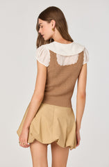 A woman is wearing a cream-colored blouse with puffed short sleeves and a large, ruffled collar. Over the blouse, she has on a fitted, textured, brown sleeveless knit top. She is also wearing beige, bubble-style shorts with a gathered, voluminous silhouette.