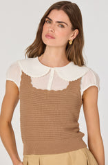 A woman is wearing a white short-sleeved blouse with a rounded collar that has scalloped edges. Over the blouse, she has on a sleeveless, light brown crochet tank top with a ribbed hem. She is also wearing beige high-waisted pants.