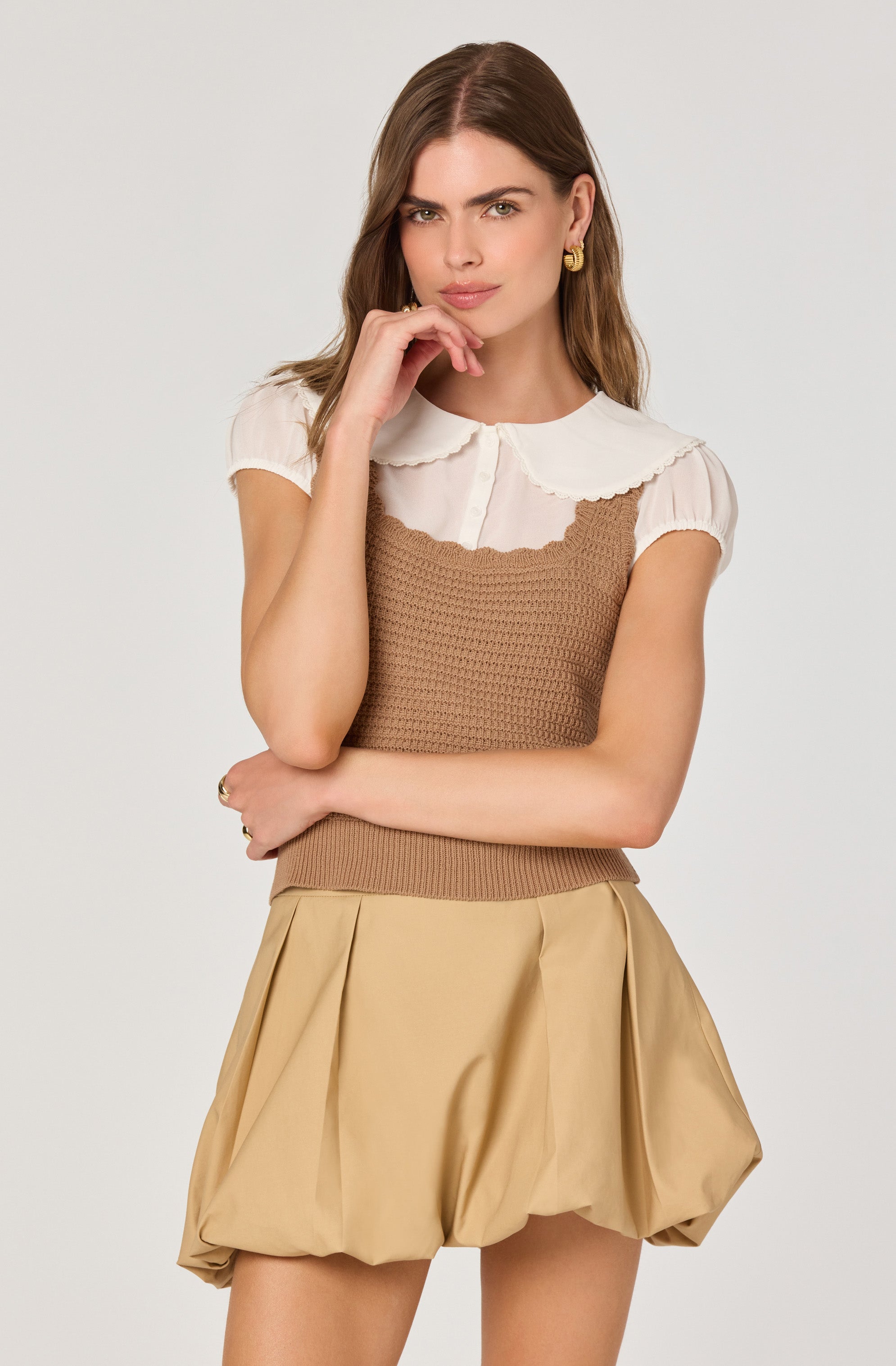 A woman is wearing a white short-sleeve blouse with a rounded, scalloped collar and a brown, textured sleeveless knit top layered over it. She pairs this with tan, voluminous, pleated shorts.