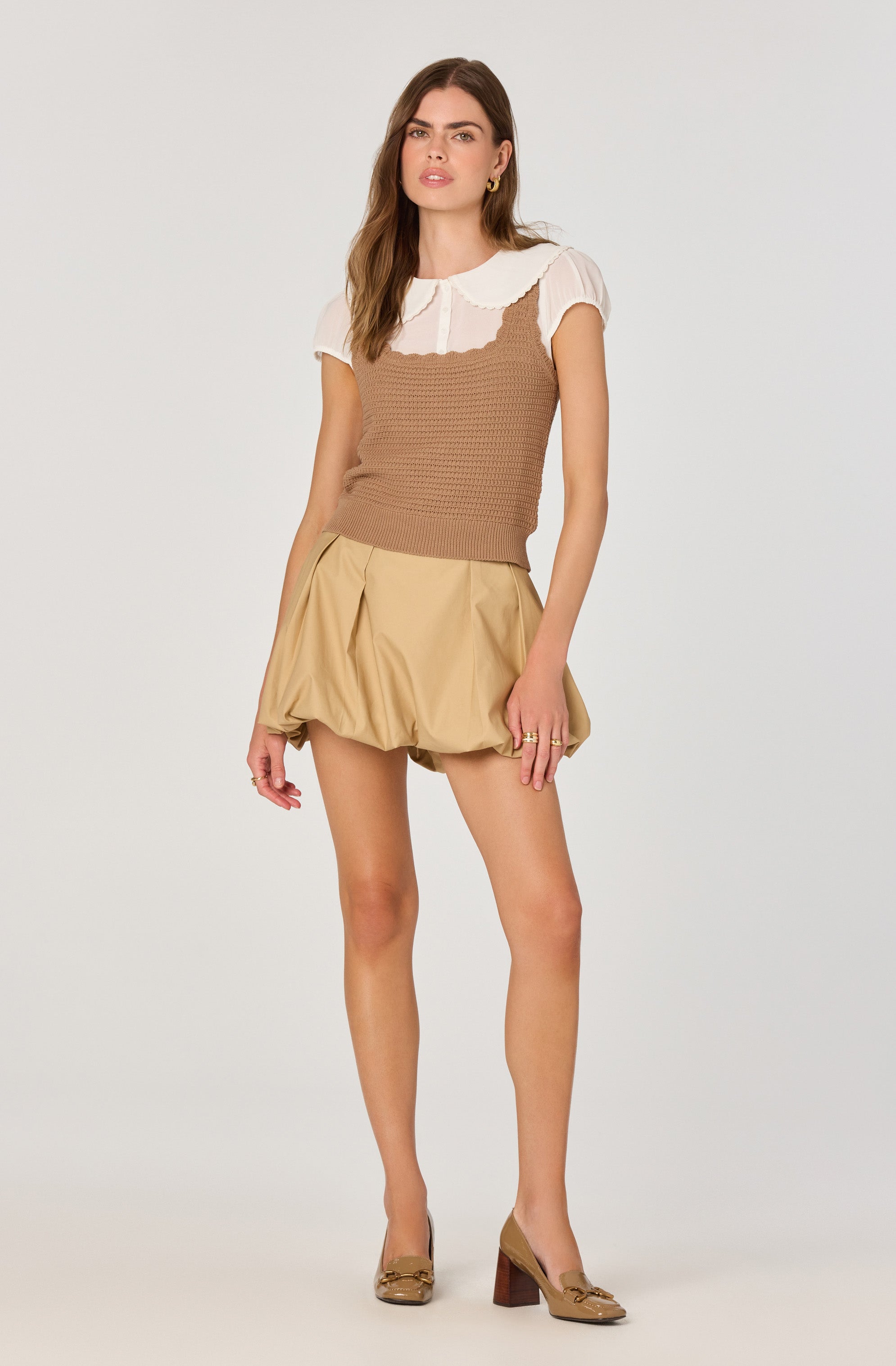The model is wearing a short-sleeved white blouse with a rounded, scalloped collar and puffed sleeves. Over the blouse, she has a sleeveless, fitted, brown knitted top. She pairs this with beige, voluminous, bubble-style shorts that have a textured, puffy appearance. She completes the outfit with beige high-heeled shoes featuring a bow detail on the toe.