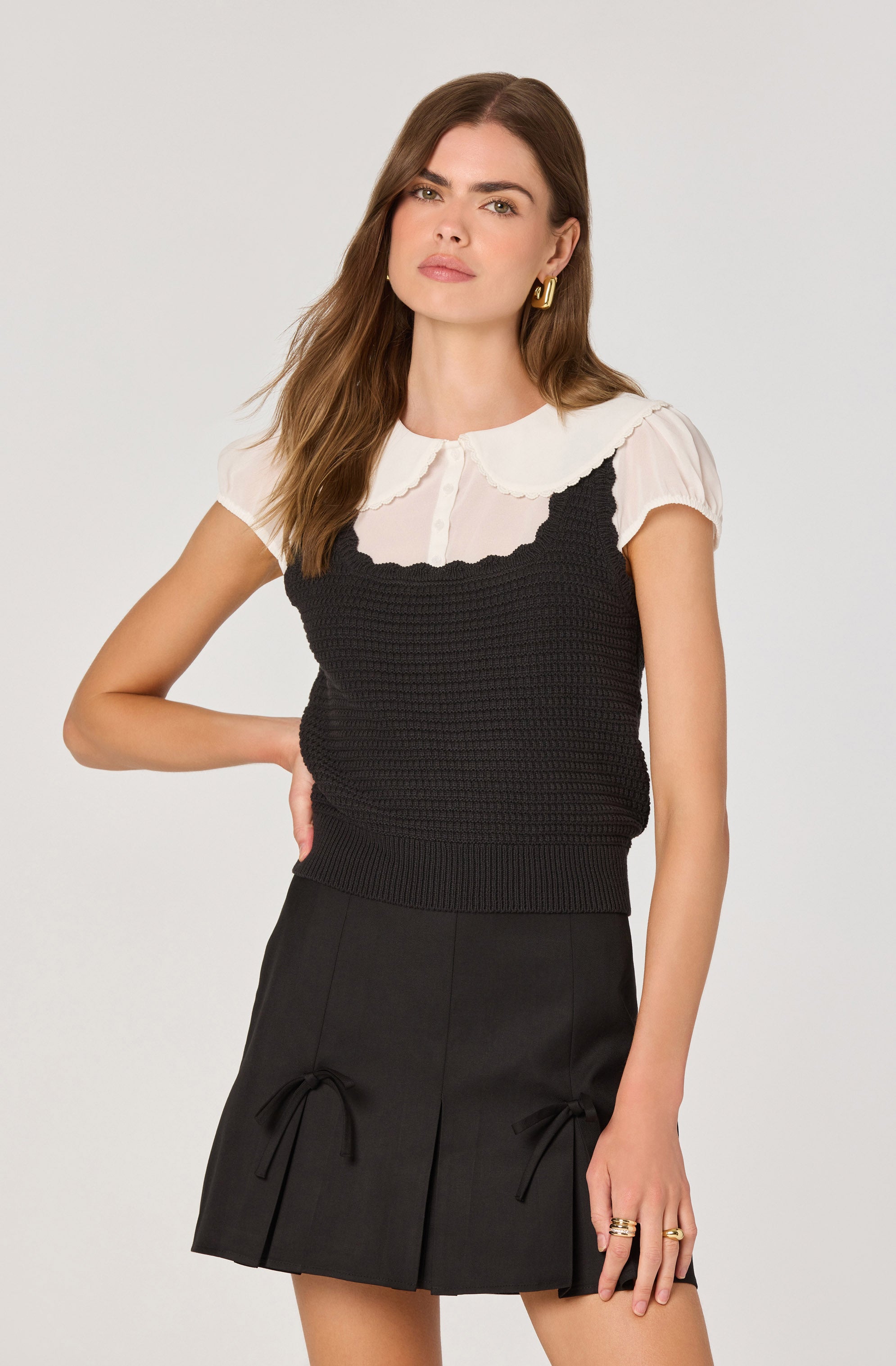 A woman is wearing a short-sleeved white blouse with a rounded collar and button-down front, layered under a black, sleeveless, textured knit top. She pairs this with a black skirt that features front pleats and decorative bow ties on each side.