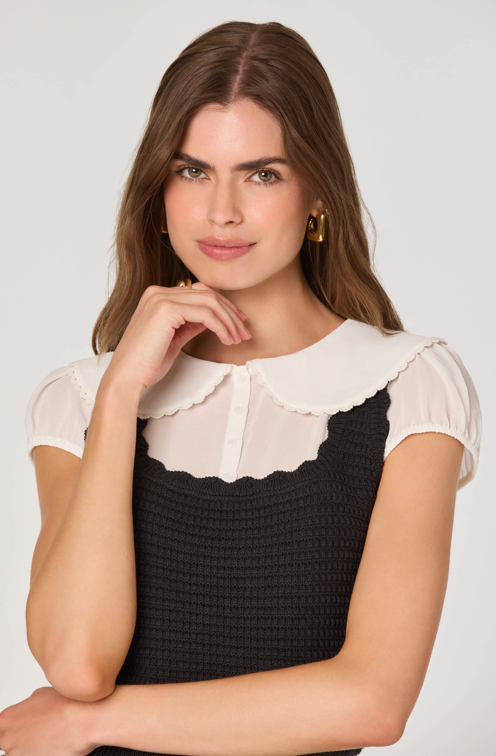 A woman is wearing a white short-sleeved blouse with a rounded collar and scalloped edges, layered under a black crocheted sleeveless top. She has gold hoop earrings and her hair is styled in loose waves. She has a confident expression with a slight smile and her hand resting near her chin.