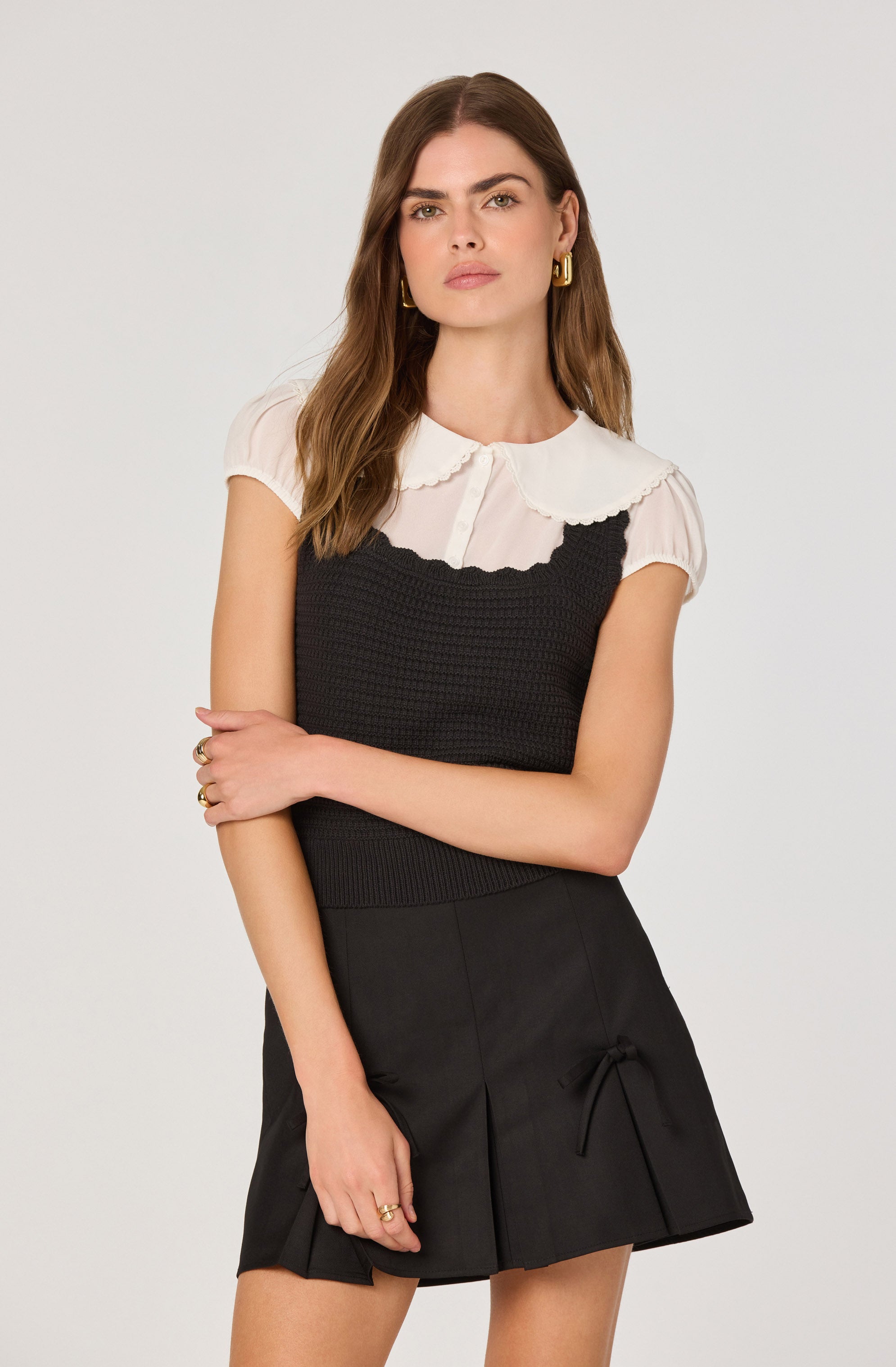 The woman is wearing a cream-colored blouse with a rounded, scalloped collar and short, puffed sleeves. Over the blouse, she has on a sleeveless black knitted top with a slightly scalloped neckline. She pairs these with a black high-waisted skirt that has pleats and a small bow detail on the side. She also wears gold hoop earrings and multiple rings.