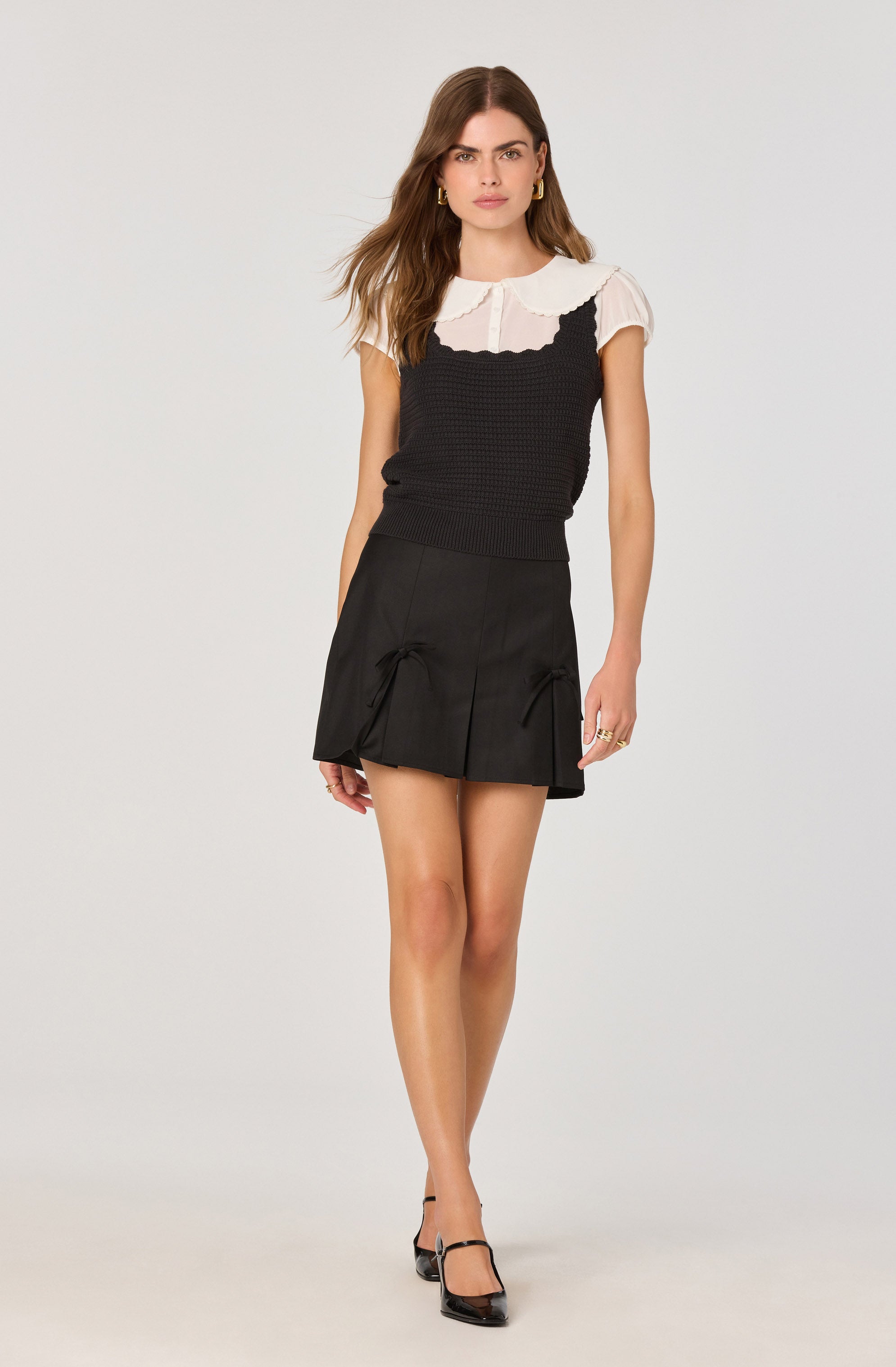 A woman is wearing a white short-sleeved blouse with a rounded collar and button detail at the front, layered under a black sleeveless textured knit top. She pairs this with a black mini skirt featuring pleats and decorative bows on the front. She completes her outfit with black high-heeled shoes with ankle straps.
