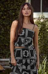 A woman with long, wavy brown hair is wearing a black two-piece outfit featuring white embroidered patterns. The top is a sleeveless, square-neck crop top with thin spaghetti straps, and the matching high-waisted pants have an elastic waistband. The embroidery consists of intricate floral and geometric designs arranged in a patchwork style. She is standing outdoors, with greenery and a window in the background.