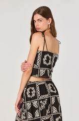 A woman is wearing a black sleeveless crop top with thin, tied shoulder straps and a matching black skirt with a white embroidered floral and geometric pattern.