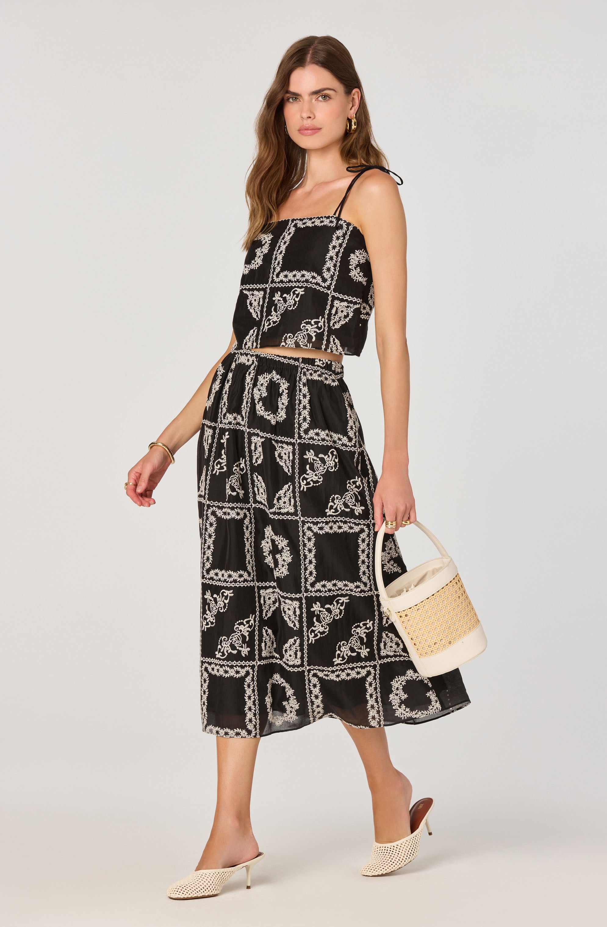 A woman is wearing a black two-piece outfit consisting of a spaghetti strap crop top and a matching midi skirt. The fabric features a white, intricate, bandana-inspired pattern arranged in a grid formation. She accessorizes with gold hoop earrings and a gold bracelet, and carries a white woven bucket bag. She is wearing open-toe, beige knit high-heeled mules.