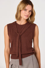 The woman is wearing a sleeveless, dark brown knitted top with a matching knitted tie at the neck. She pairs it with high-waisted, light-colored checkered trousers.