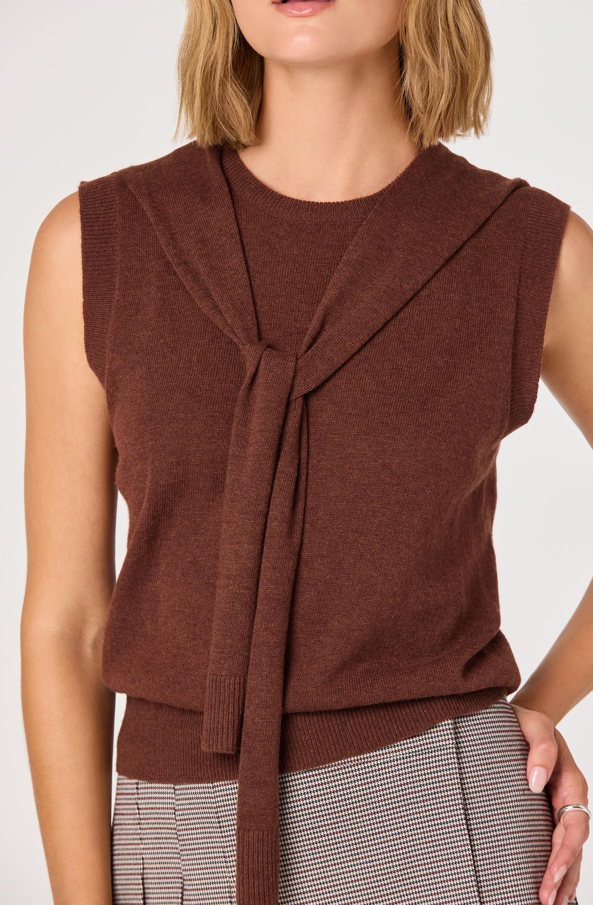 A sleeveless, knit sweater vest in a warm brown color featuring a decorative tied detail at the front. The vest has a round neckline and a ribbed hem at the waist.