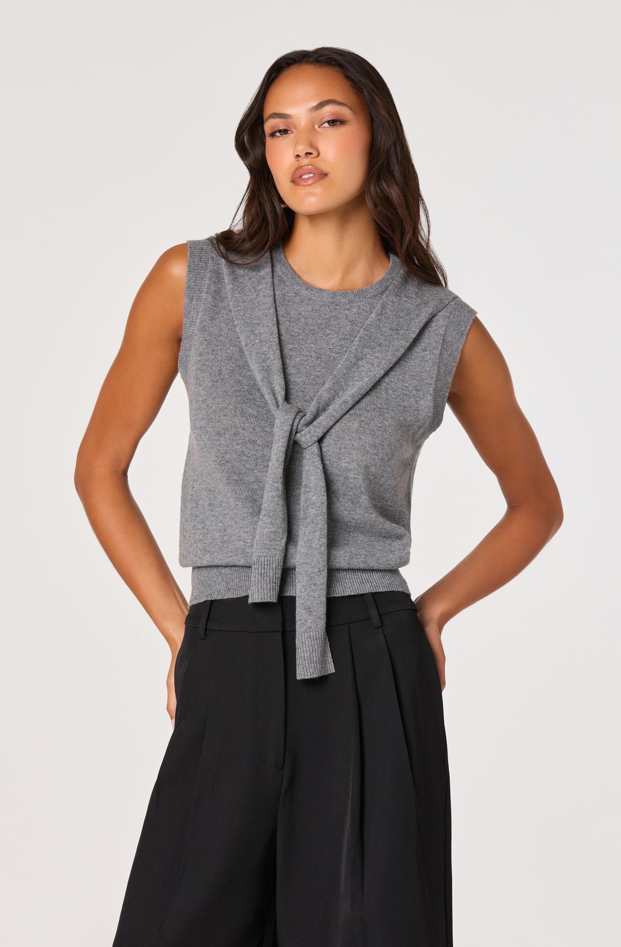 A woman is wearing a sleeveless gray knit top with a matching gray cardigan tied around her shoulders. She is also wearing high-waisted black wide-leg trousers with pleats.